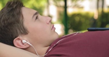 Parktaki bankta uzanmış, kulaklıklı genç bir çocuğun portresi. Ergen kulaklıkla müzik dinliyor. Genç yakışıklı adam kulaklıkla açık havada müzik dinliyor.. 