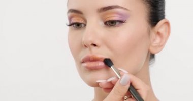 Makyöz dudaklarına parlak bir ruj sürüyor. Makyaj yapıyorum. Ruju fırçayla dudaklara sürten bir kızın yüzünü yakından çek. Profesyonel model. Gerçek zamanlı 59.94 fps. Profesyonel ışık