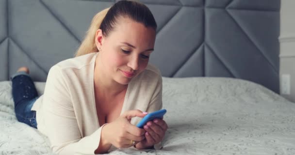 집에서 전화를 사용하는 스마트폰으로 메시지를 입력하는 성인아름다운 미소짓는 여자는 침대에 — 비디오