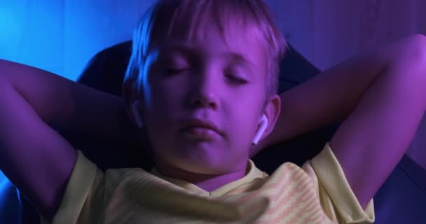 男孩在耳机里听音乐 可爱的男孩躺在沙发上 轻松地听着音乐 晚上孩子们在他的房间休息 8岁儿童的夜间闲暇 男孩对着音乐摇头 — 图库视频影像