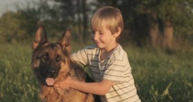 Doğada köpeği olan bir çocuk. 7 yaşında köpek besleyen bir çocuk Alman çoban köpeği besliyor. Sahada köpekle gülümseyen mutlu çocuk. Küçük çocuk çayırda evcil hayvanıyla oynuyor. Yavaş çekim. 