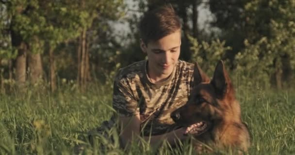 有狗在大自然中的青少年 15岁的男孩爱抚一只德国牧羊犬 和狗一起在球场上快乐地微笑着的少年 白人在草地上和他的宠物玩耍 — 图库视频影像