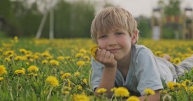 Çayırda yatan ve bakışları olan beyaz bir çocuk. Mutlu çocuk papatya tarlasında yatıyor, yakından bakıyor. Çayırdaki sarı saçlı çocuğun sevimli gülümseyen yüzü. Mutluluk ve çocukluk. Yavaş çekim