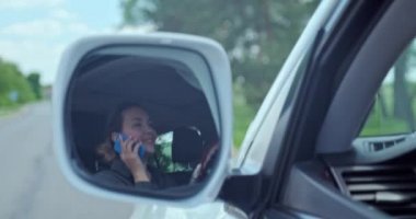 Beyaz yetişkin kız araba sürerken cep telefonuyla konuşur. Arabayı kadın kullanıyor. Arabadaki mutlu kadın telefonda konuşuyor. Araba aynasında yansıyan yansıma araba kullanan ve telefonla arayan kadının yansıması..  