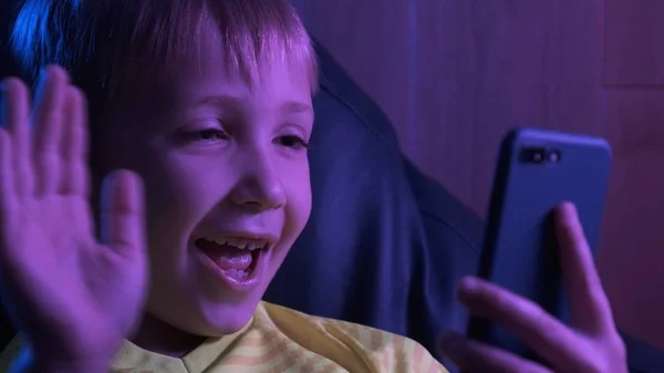 微笑的男孩看着屏幕上的智能手机，向他的飞蛾挥挥手 图库图片