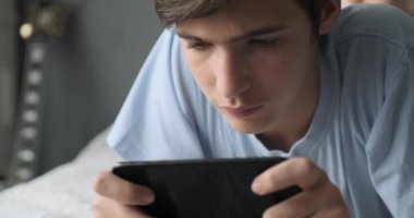 Ergen, akıllı telefonla, evde oyunlar oynuyor. Yakışıklı çocuk koltukta uzanarak cep telefonu kullanıyor. Oyuncu cep telefonuyla oynar. Yaşam tarzı konsepti. Gerçek zamanlı görüntüler