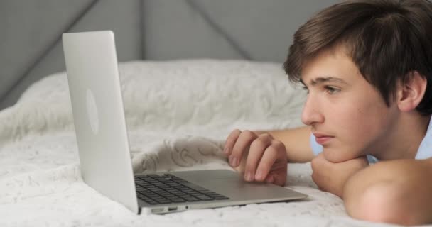 若い男は自宅のベッドに横たわってノートパソコンで動作します 昼食の時間にコンピュータを使用して10代の子供 10代は午後にコンピュータのキーボードを入力しています リアルタイムだ4K映像 — ストック動画