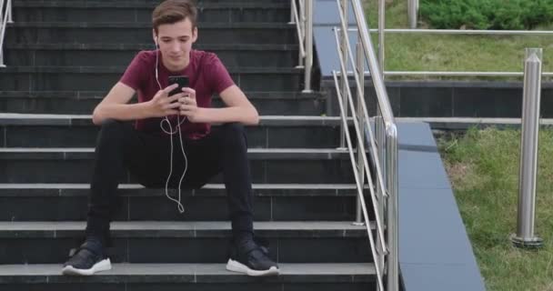 在停车场用手机的青少年 在户外用手机微笑的年轻人 快乐的男孩少年与手机 城市场景 帅哥用的是智能手机 — 图库视频影像