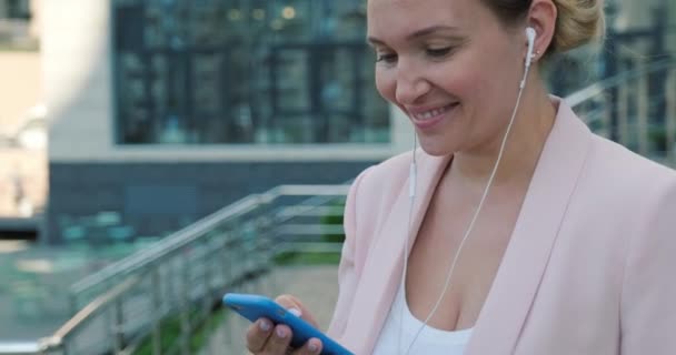 女人听耳机里的音乐 笑着的成年女人在户外用耳机听音乐 一个戴耳机的快乐女士的画像 城市场景 — 图库视频影像