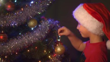 Noel Baba şapkalı çocuk Noel ağacına Noel oyuncakları asıyor. Mutlu 8 yaşındaki çocuk yeni yıla hazırlanıyor. Parlak oyuncaklar ve çelenklerle Noel ağacında gülümseyen bir çocuğun 4K videosu.