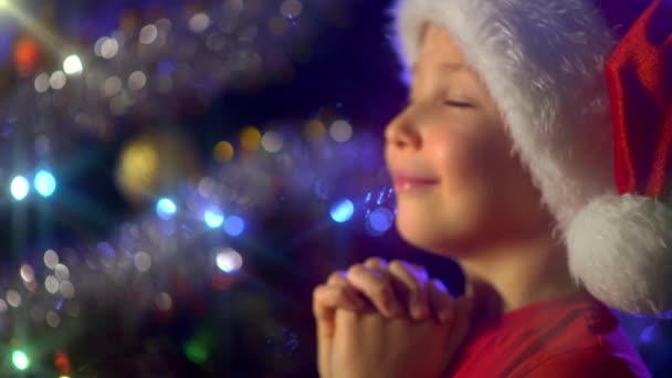 一个笑着的男孩在圣诞树旁祈祷圣诞节礼物的4K视频 戴着圣诞礼帽的男孩正在梦想着圣诞节的圣诞礼物 快乐的8岁小孩在为新年做准备 — 图库视频影像