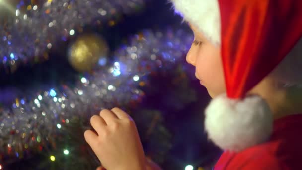 笑顔の男の子の4Kビデオはクリスマスプレゼントについてクリスマスツリーの近くで祈っています サンタの帽子の男の子はクリスマスにクリスマスの贈り物を夢見ています 幸せな8歳の子供は新年の準備をしています — ストック動画