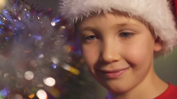 Garçon dans un chapeau de Père Noël accroche des jouets de Noël sur le sapin de Noël. Joyeux enfant se prépare pour la nouvelle année. Vidéo 4K d'un garçon souriant au sapin de Noël avec des jouets lumineux et des guirlandes. Gros plan sur le visage 