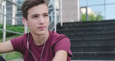 Ergen kulaklıkla müzik dinliyor. Gülümseyen genç yakışıklı adam açık havada, kulaklıkla müzik dinliyor. Kulaklık takmış mutlu bir gencin portresi. Şehir manzarası. 4 bin. Gerçek zamanlı..