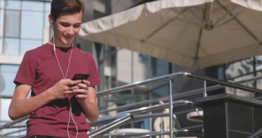 Teen using mobile phone in park. Smiling young man using cell phone, outdoors. Happy boy teenager with mobile phone. Urban scene. Handsome guy uses smartphone. Slow motion