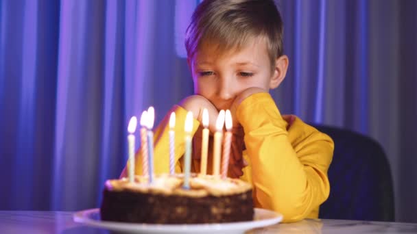 悲しい少年は誕生日にろうそくを灯したケーキを探します 誕生日に白人の子供を動揺させた お子様一人でお菓子の前にろうそくを立てて座ります 不幸な白い子供は誕生日ケーキを見て — ストック動画