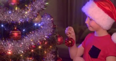 Noel Baba şapkalı çocuk Noel ağacına Noel oyuncakları asıyor. Mutlu 8 yaşındaki çocuk yeni yıla hazırlanıyor. Parlak oyuncaklar ve çelenklerle Noel ağacında gülümseyen bir çocuğun 4K videosu.