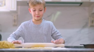 Merdaneli hamuru yuvarlayan çocuk, yakın görüş. Çocuk hamuru mutfak masasının üzerine atıyor. 8 yaşındaki çocuk evde yemek hazırlıyor. Çocuk merdaneyle hamuru yuvarlıyor. Yavaş çekim 4k görüntü 