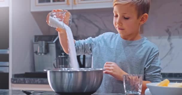 白い男の子はクッキーを調理するためにガラスからボウルに砂糖を注ぎ キッチンで子供の料理を閉じます スローモーション4K映像 — ストック動画
