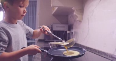 Mutfakta yemek pişiren 8 yaşındaki bir çocuk. Çocuk kepçeden tavaya krep kızartmak için karışımı dolduruyor. Çocuk krep karışımını çırpıcıyla çırpıyor. Ağır çekim. Çocukluk kavramı
