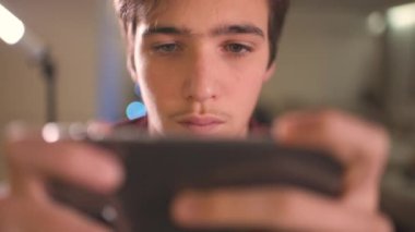 Evde akıllı telefonlu bir gencin yüzünü kapat. Kafkasyalı genç adam sosyal ağda cep telefonu kullanarak zaman geçiriyor. Beyaz genç adam oyun için cep telefonu kullanıyor. Yaşam tarzı kavramı. 