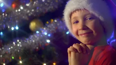 Gülümseyen bir çocuğun Noel ağacının yanında Noel hediyeleri için dua ettiği 4K videosu. Noel Baba şapkalı çocuk Noel hediyeleri hayal ediyor. Mutlu 8 yaşındaki çocuk yeni yıla hazırlanıyor.. 