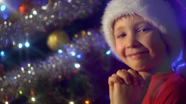 笑顔の男の子の4K動画はクリスマスツリーの近くでクリスマスプレゼントについて祈っています サンタの帽子の男の子はクリスマスにクリスマスの贈り物を夢見ています 幸せな8歳の子供は新年の準備をしています — ストック動画
