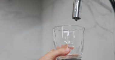 Çocuğun eli, mutfak musluğunun yanında damlayan suyla şeffaf bir bardak tutuyor. Çocuğun eli temiz bir bardak tutuyor ve onu temiz suyla dolduruyor. Küresel tatlı su sıkıntısı sorunu