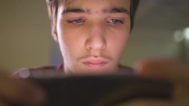 自宅で10代の男の子の顔をスマートフォンで閉じます 白人の10代の男は携帯電話を使ってソーシャルネットワークに時間を費やしています 白人の若者が携帯電話を使ってゲームをしてる ライフスタイルコンセプト — ストック動画