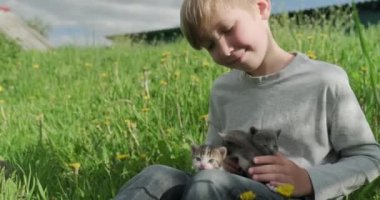 Çocuk küçük kedileri elinde tutuyor. Ellerinde kedi yavrusu olan mutlu çocuk çimlerin üzerinde oturuyor. Evin dışında elinde üç evcil hayvanla çimenlerin üzerinde oturan sevimli bir bebek. Gerçek zamanlı.