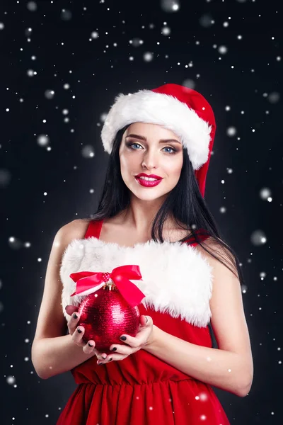 Fille souriante tenant une boule d'arbre de Noël rouge. Des femmes en robe et chapeau de Père Noël. Aide du Père Noël. Jolie fille heureuse dans le chapeau du Père Noël avec des lèvres rouges posant pour la caméra sur fond noir de fête de Noël . — Photo