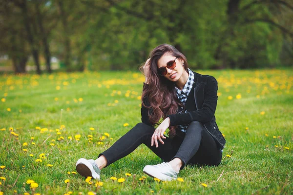 Chica joven con estilo en una camisa a cuadros y gafas de sol sentado en la hierba verde en la primavera — Foto de Stock