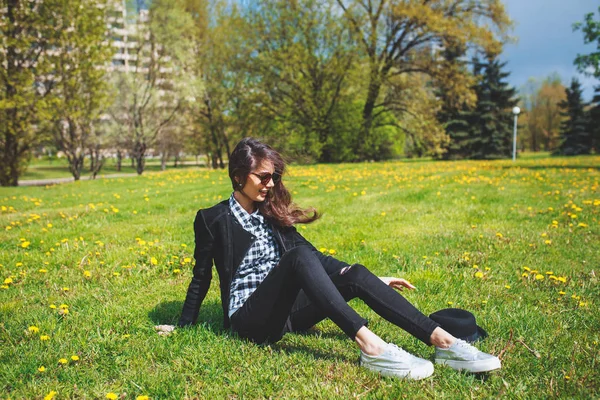 Стильна молода дівчина в плетеній сорочці і сонцезахисних окулярах, що сидять на зеленій траві навесні — стокове фото