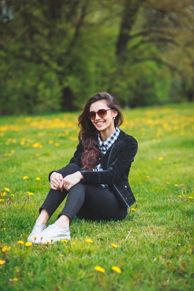 Chica joven con estilo en una camisa a cuadros y gafas de sol sentado en la hierba verde en la primavera — Foto de Stock