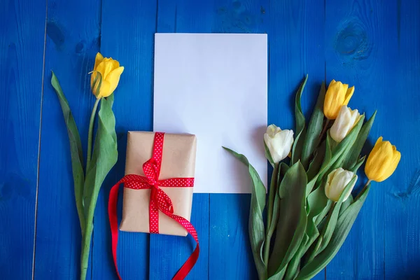 Lente tulp bloemen, doos en papieren kaartje op blauwe houten tafel van bovenaf in plat lag stijl. — Stockfoto