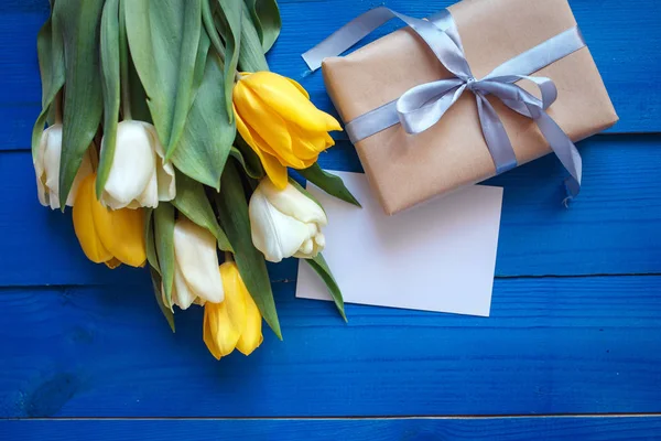 Lente tulp bloemen, doos en papieren kaartje op blauwe houten tafel van bovenaf in plat lag stijl. — Stockfoto