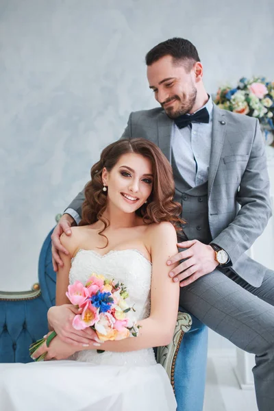 Bruid in mooie jurk en bruidegom in grijs pak op sofa binnenshuis in witte studio interieur zoals om thuis te zitten. Trendy bruiloft stijl — Stockfoto