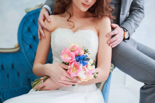 Bruid in mooie jurk en bruidegom in grijs pak op sofa binnenshuis in witte studio interieur zoals om thuis te zitten. Trendy bruiloft stijl — Stockfoto