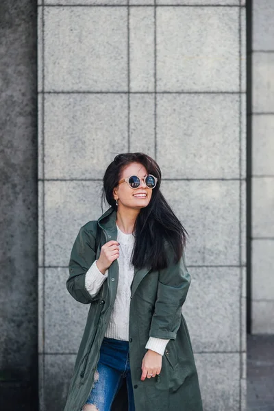 Fashion closeup portrait of nice pretty young hipster woman posing in sunglasses Outdoor .Brunette happy girl in green raincoat and pink sneakers walks the street of the city. — Stock Photo, Image
