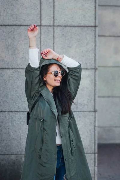 Mode närbild porträtt av trevlig ganska ung hipster kvinna poserar i solglasögon utomhus. Brunett glad tjej i grön regnrock och rosa sneakers går gatan av staden. — Stockfoto