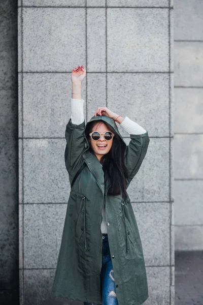 Fashion closeup portrait of nice pretty young hipster woman posing in sunglasses Outdoor .Brunette happy girl in green raincoat and pink sneakers walks the street of the city. — Stock Photo, Image