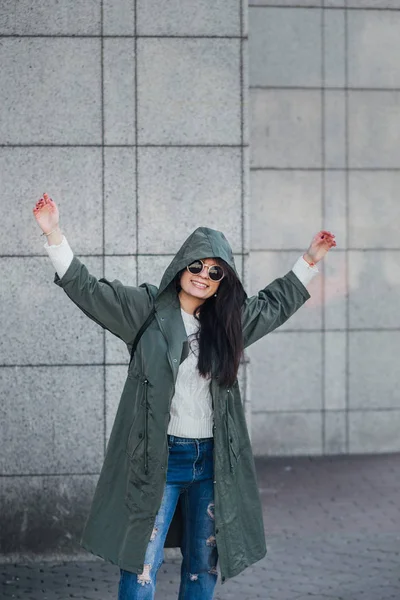 Mode närbild porträtt av trevlig ganska ung hipster kvinna poserar i solglasögon utomhus. Brunett glad tjej i grön regnrock och rosa sneakers går gatan av staden. — Stockfoto