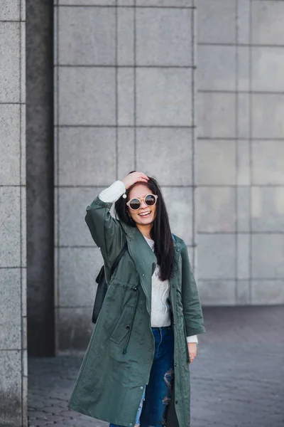 Retrato de cerca de la moda de bonita mujer hipster bastante joven posando en gafas de sol al aire libre .Brunette chica feliz en impermeable verde y zapatillas de deporte de color rosa pasea por la calle de la ciudad . — Foto de Stock