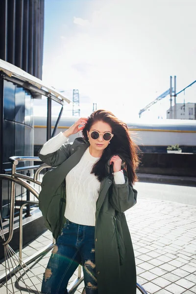 Fashion closeup portrait of nice pretty young hipster woman posing in sunglasses Outdoor .Brunette happy girl in green raincoat and pink sneakers walks the street of the city. — Stock Photo, Image