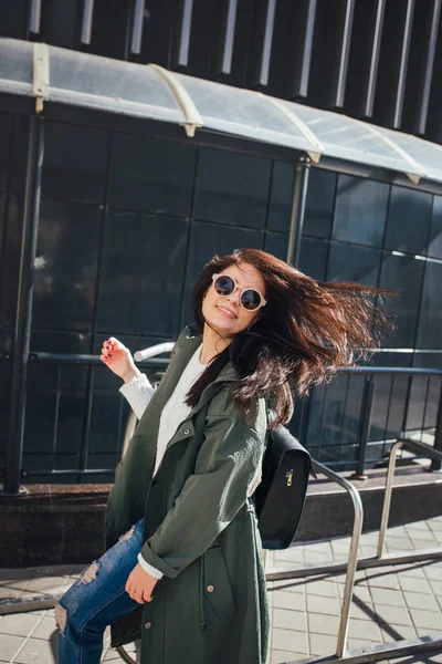 Retrato de cerca de la moda de bonita mujer hipster bastante joven posando en gafas de sol al aire libre .Brunette chica feliz en impermeable verde y zapatillas de deporte de color rosa pasea por la calle de la ciudad . — Foto de Stock