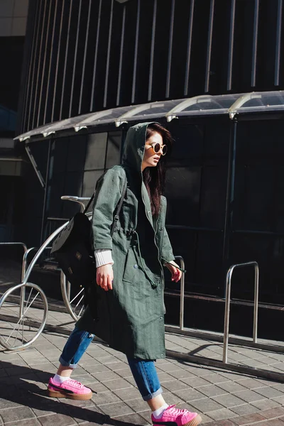 Retrato de cerca de la moda de bonita mujer hipster bastante joven posando en gafas de sol al aire libre .Brunette chica feliz en impermeable verde y zapatillas de deporte de color rosa pasea por la calle de la ciudad . — Foto de Stock