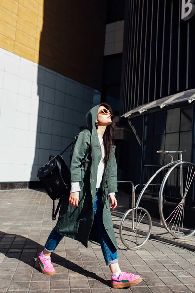 Retrato de cerca de la moda de bonita mujer hipster bastante joven posando en gafas de sol al aire libre .Brunette chica feliz en impermeable verde y zapatillas de deporte de color rosa pasea por la calle de la ciudad . — Foto de Stock