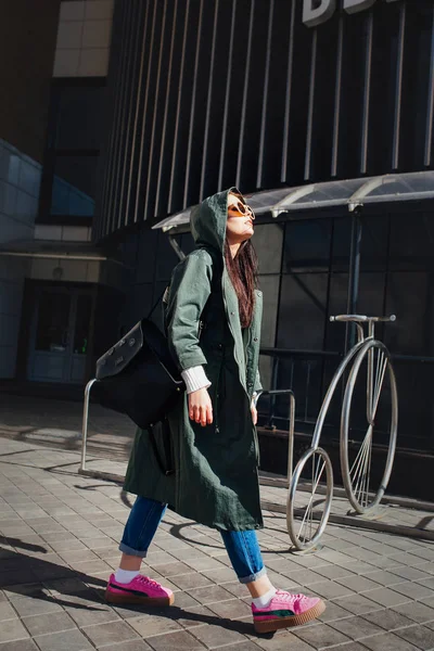 Retrato de cerca de la moda de bonita mujer hipster bastante joven posando en gafas de sol al aire libre .Brunette chica feliz en impermeable verde y zapatillas de deporte de color rosa pasea por la calle de la ciudad . — Foto de Stock