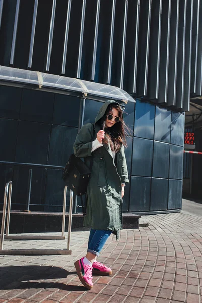 Retrato de cerca de la moda de bonita mujer hipster bastante joven posando en gafas de sol al aire libre .Brunette chica feliz en impermeable verde y zapatillas de deporte de color rosa pasea por la calle de la ciudad . — Foto de Stock