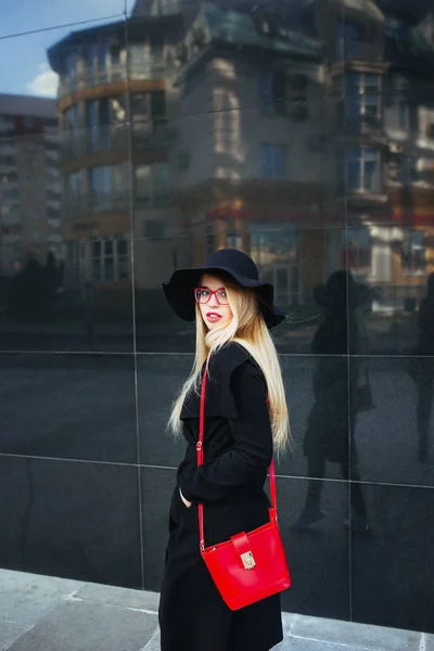 Bella giovane donna in un cappotto nero e cappello, indossando occhiali rossi e una borsa rossa che cammina in città. Riflessione della città nella finestra — Foto Stock